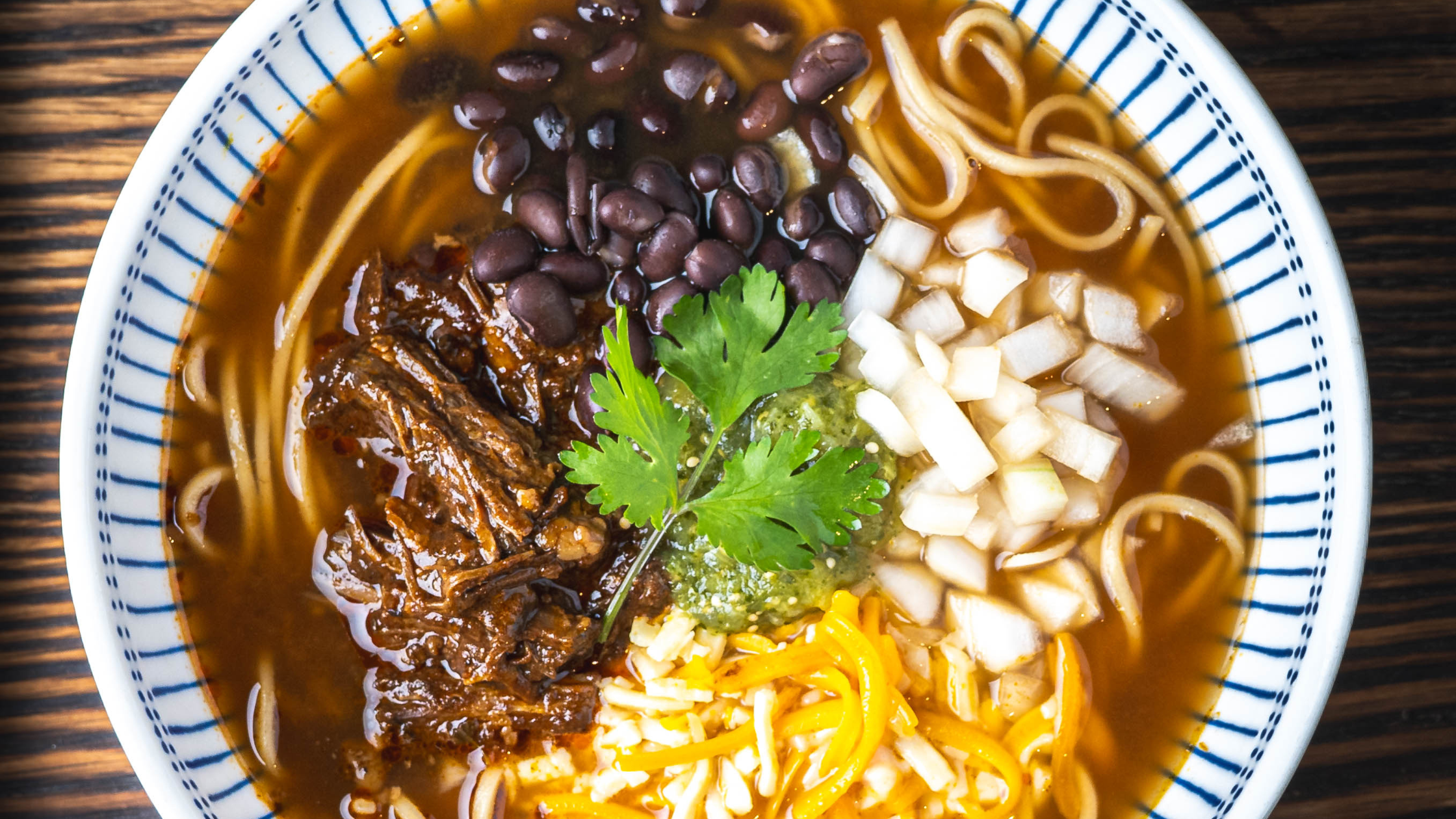 Topdown view of finished Beef Birrira Frozen Ramen in a bowl with masa noodles, beef birria, cheese, onions, and black beans.