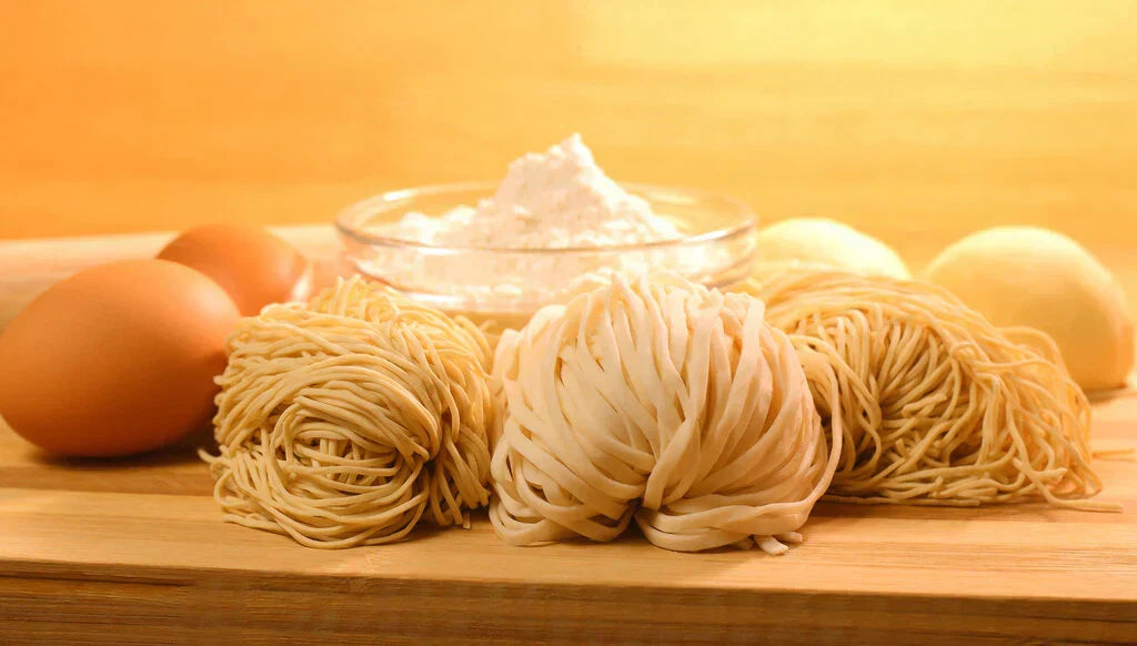 Bundles of ramen and pasta noodles next to eggs and a bowl of flour.