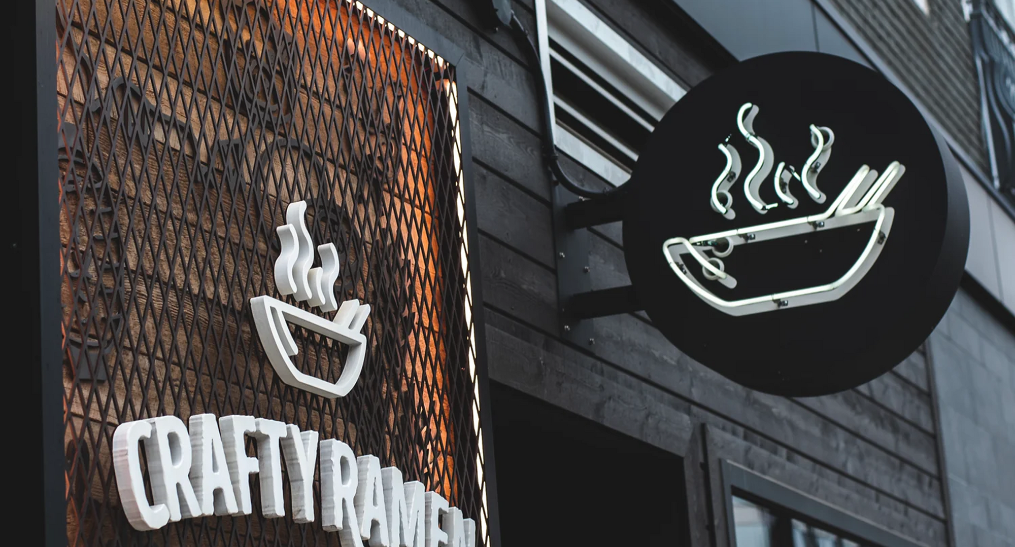 Exterior storefront signage for Crafty Ramen Noodle Shop featuring white neon bowl icons with steam wisps on orange mesh and black backgrounds, with a yellow pendant light fixture above and dark storefront windows.