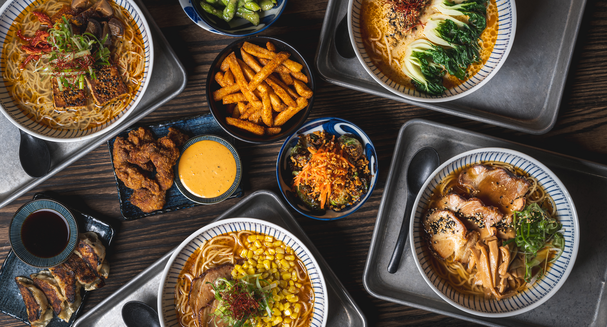 An overhead flat lay of various ramen bowls served with sides including fried chicken, fries, edamame, corn, and dipping sauce on a dark wooden table.