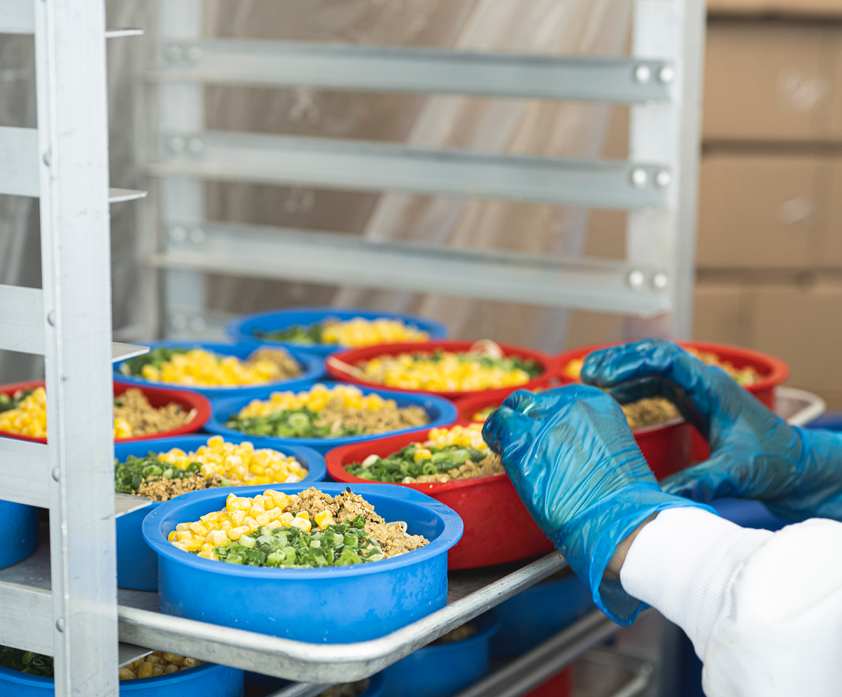 An overhead view of multiple blue and red plastic containers filled with ramen toppings including corn, green onions, and ground meat on metal trays in a food preparation facility with white shelving.