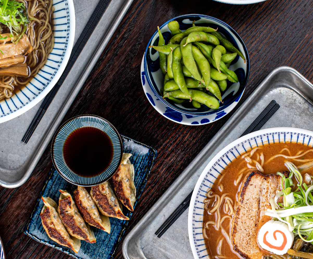 A dining table with ramen bowls, steamed dumplings, edamame, and soy dipping sauce arranged with chopsticks on a dark wooden surface.