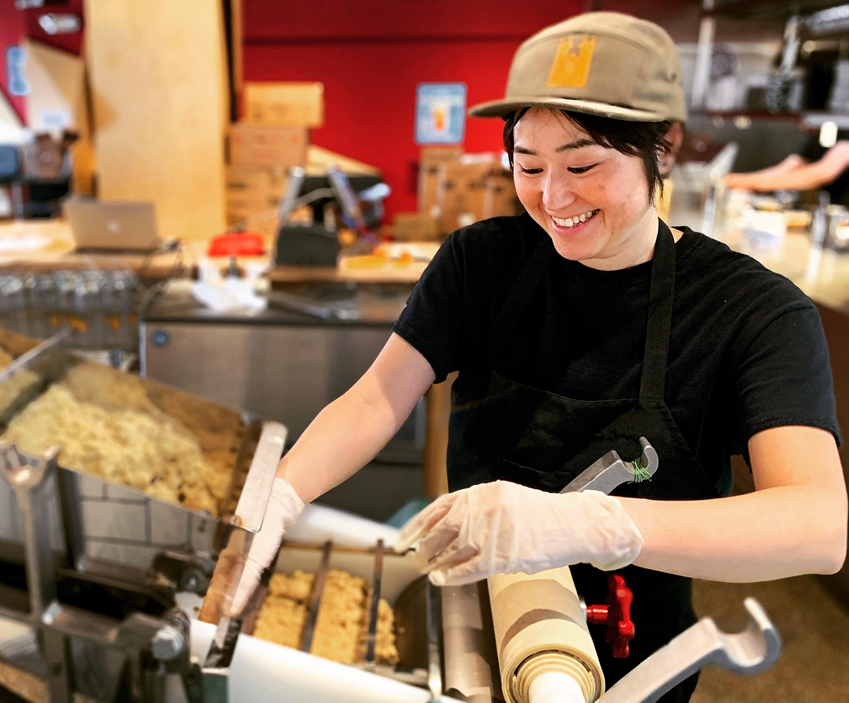 A smiling employee wearing a black shirt and beige cap working at a Crafty Ramen facility, preparing noodles with cooking equipment visible in the background.