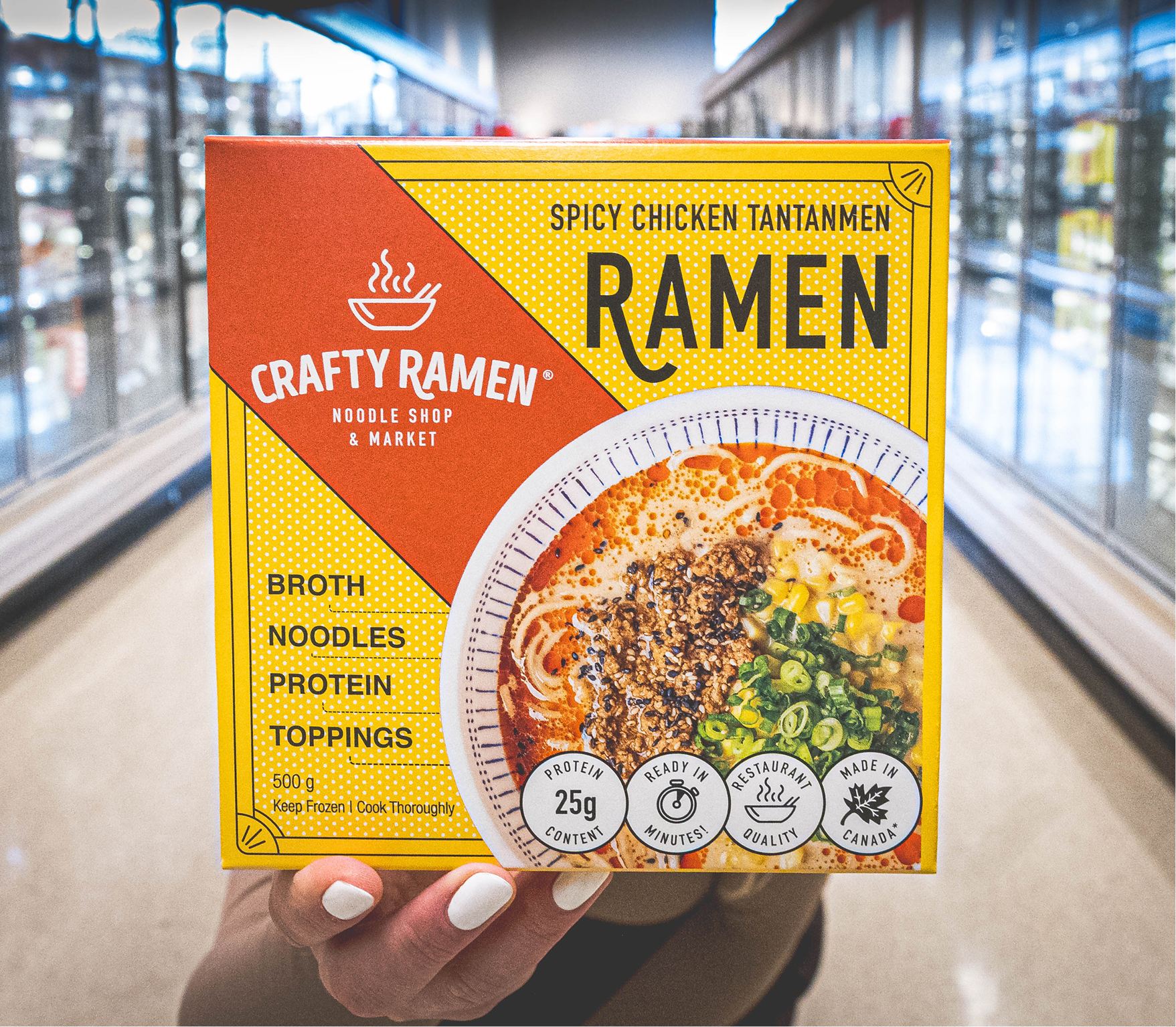 A hand holding a box of Crafty Ramen Spicy Chicken Tantanmen with red and yellow packaging and product imagery, taken in a grocery store aisle.