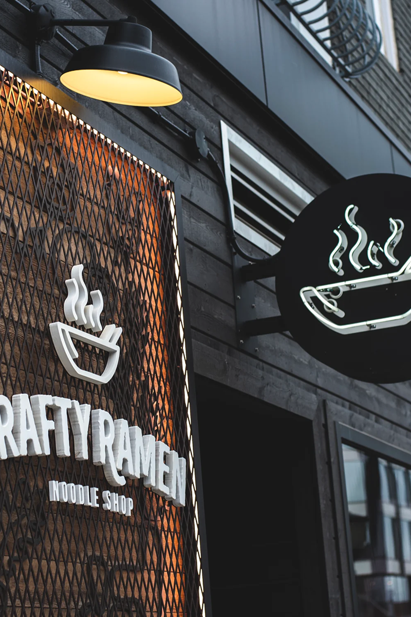 Exterior storefront signage for Crafty Ramen Noodle Shop featuring white neon bowl icons with steam wisps on orange mesh and black backgrounds, with a yellow pendant light fixture above and dark storefront windows.