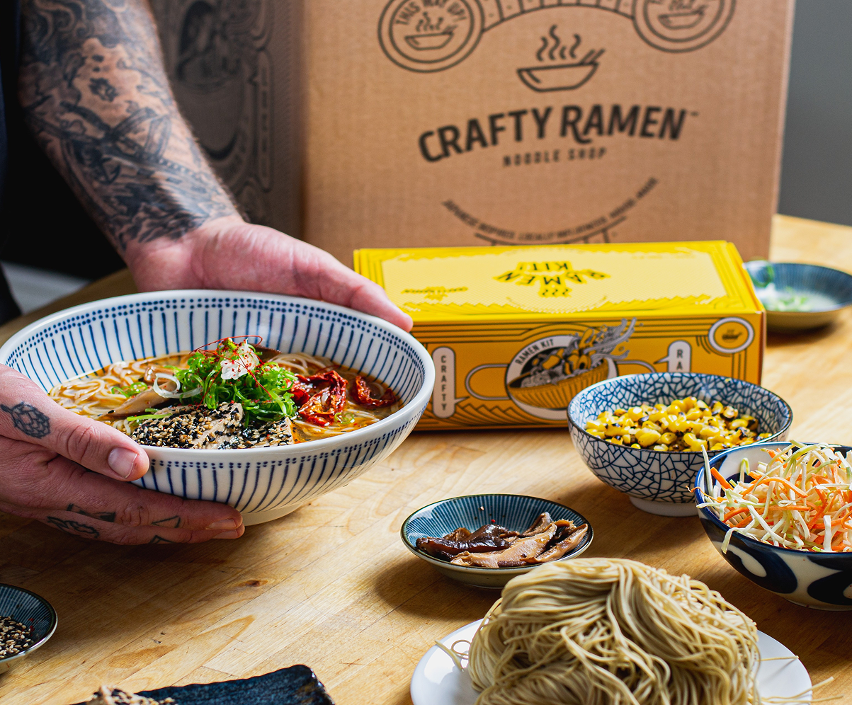 A hand holding a bowl of spicy ramen with red chili peppers, black sesame seeds, green onions, and garnish, surrounded by small bowls of toppings and a Crafty Ramen box.