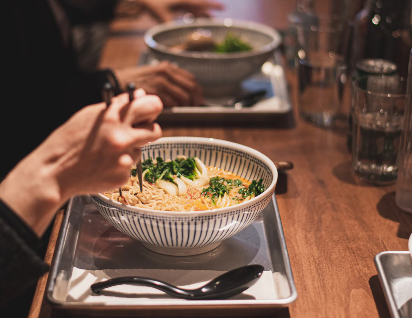 A person eating ramen at a restaurant table with chopsticks, holding a blue and white striped bowl of noodles in broth garnished with green onions.