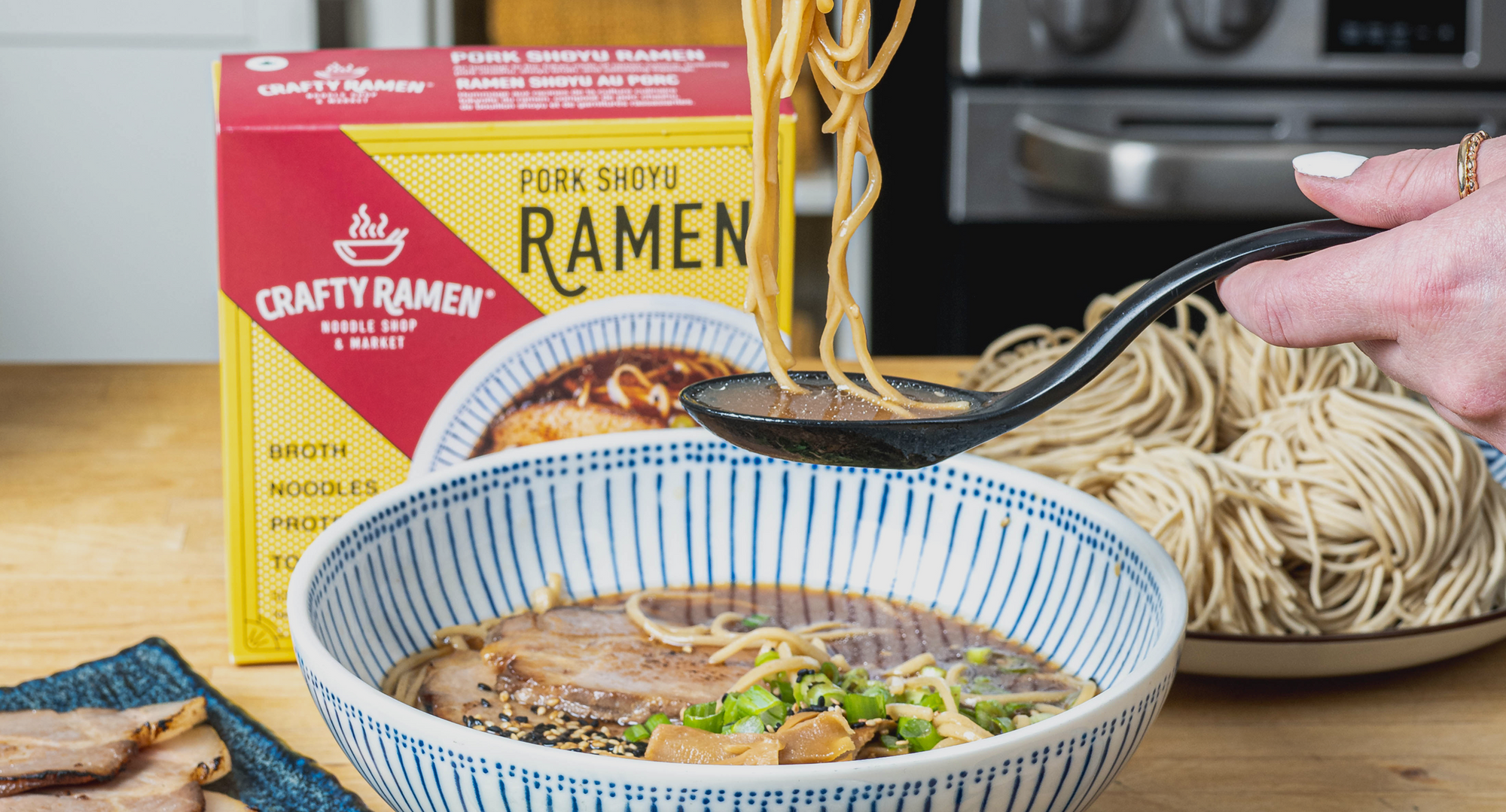 A hand holding chopsticks lifts ramen noodles from a blue and white striped bowl containing pork shoyu ramen with broth, noodles, sliced pork, green onions, and sesame seeds, with a Crafty Ramen Pork Shoyu box visible in the background.