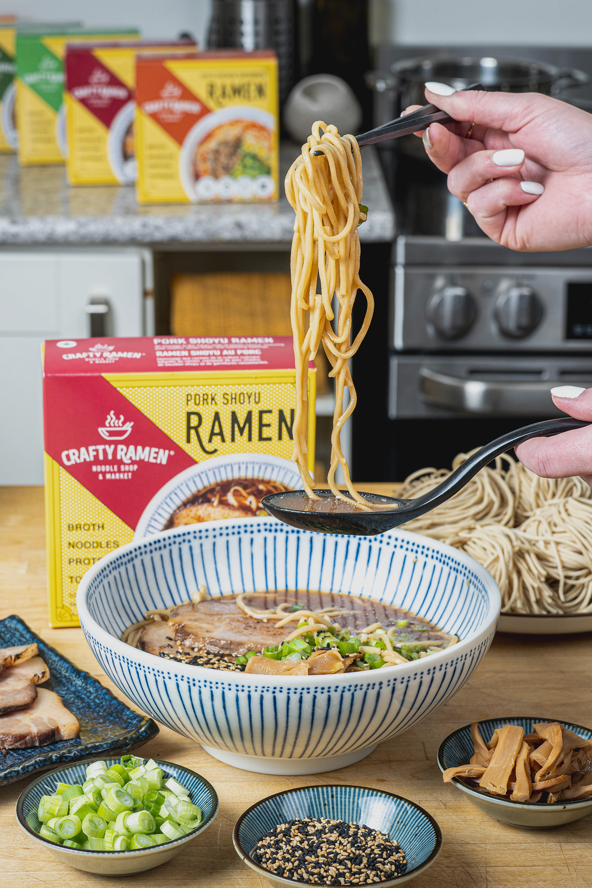 Chopsticks lifting a bundle of golden ramen noodles from a bowl of broth, with a Crafty Ramen Pork Shoyu box and multiple ramen packages visible in the blurred background.
