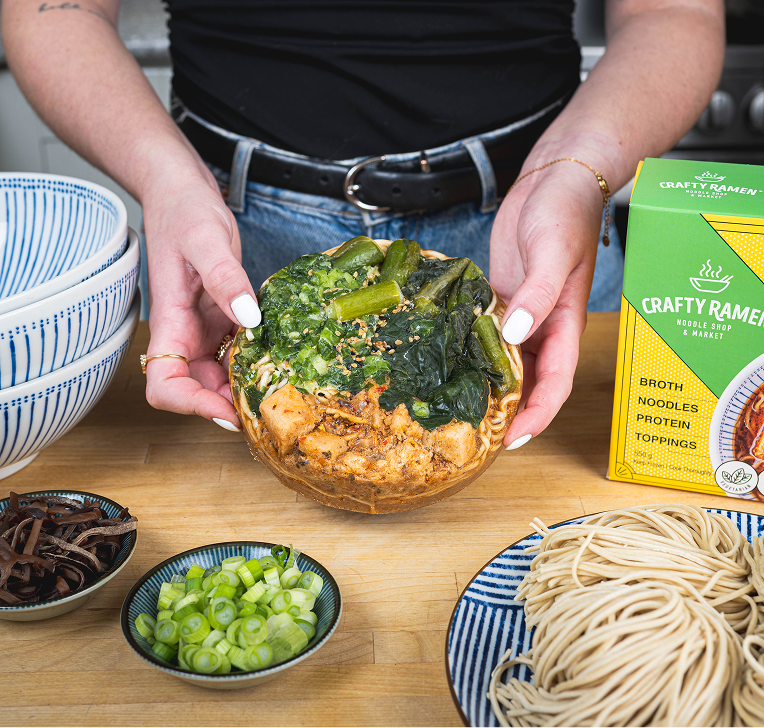 Hands holding an open green packet containing what appears to be ramen noodles and seasonings, with ramen bowls, a blue spice container, and green onion garnish visible on the wooden table below.