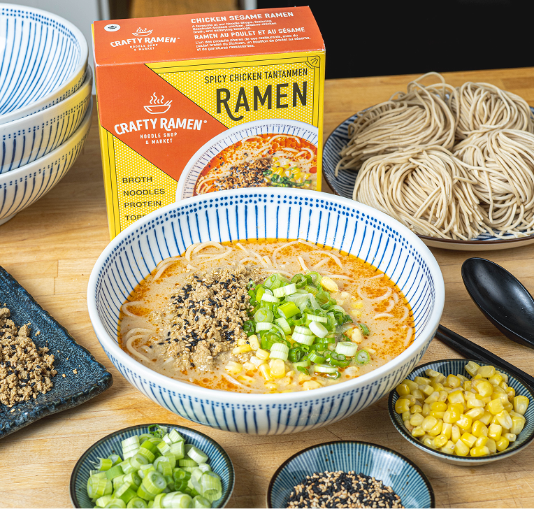 An overhead view of a bowl of Spicy Chicken Tantanmen ramen garnished with ground meat, sesame seeds, and green onions, next to an orange Crafty Ramen box with small bowls of toppings nearby.