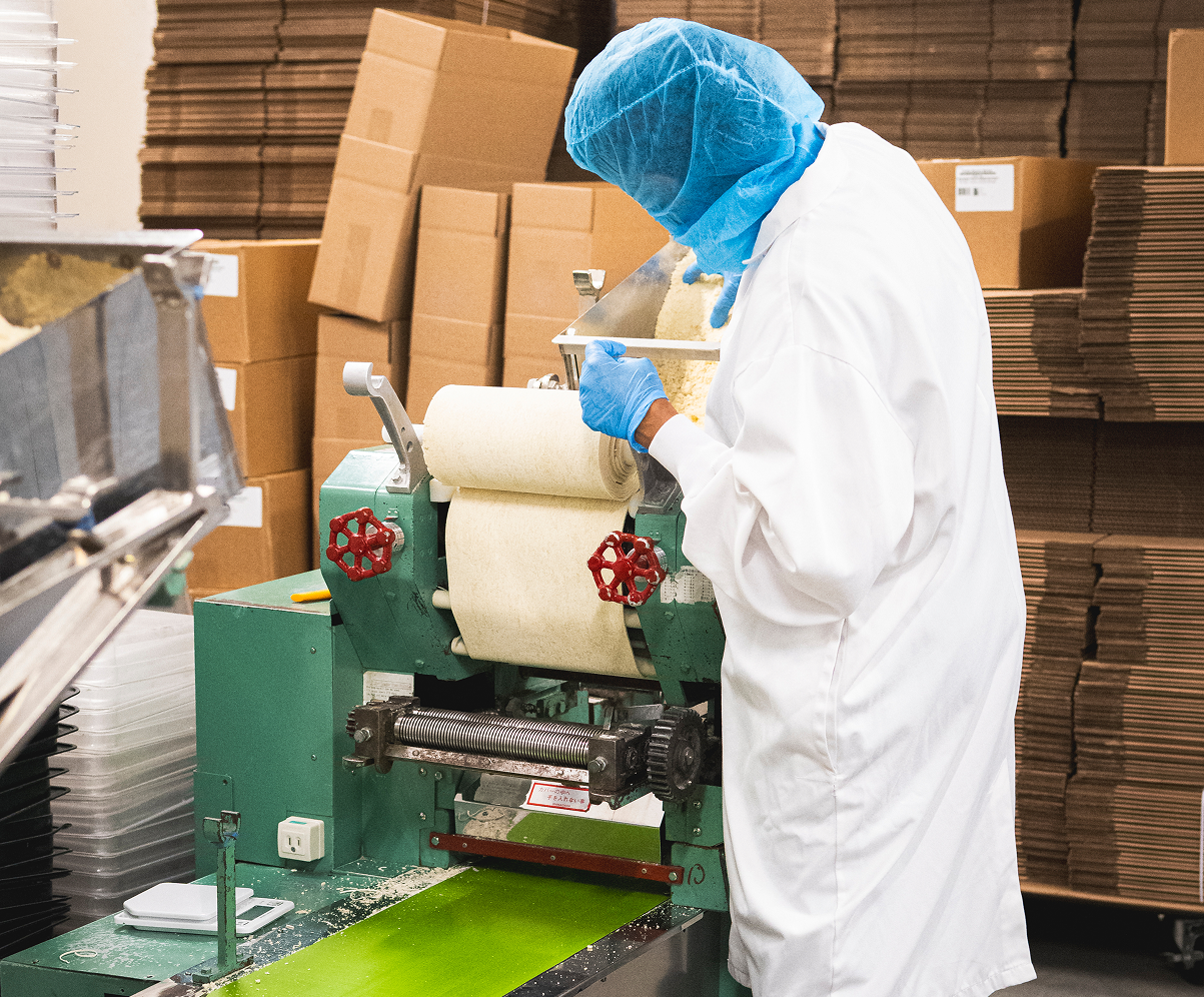A worker in white coat and blue hairnet and gloves operating a large industrial noodle-making machine in a production facility with cardboard boxes in the background.