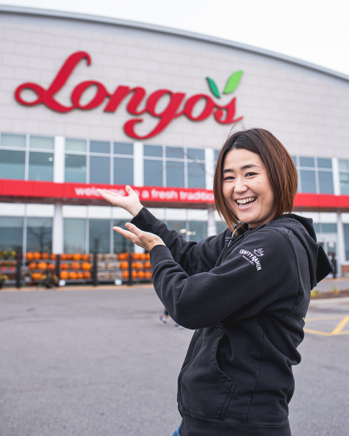 Miki smiling and gesturing toward a Longo's building, wearing a black Crafty Ramen hoodie with a white logo on the shoulder.