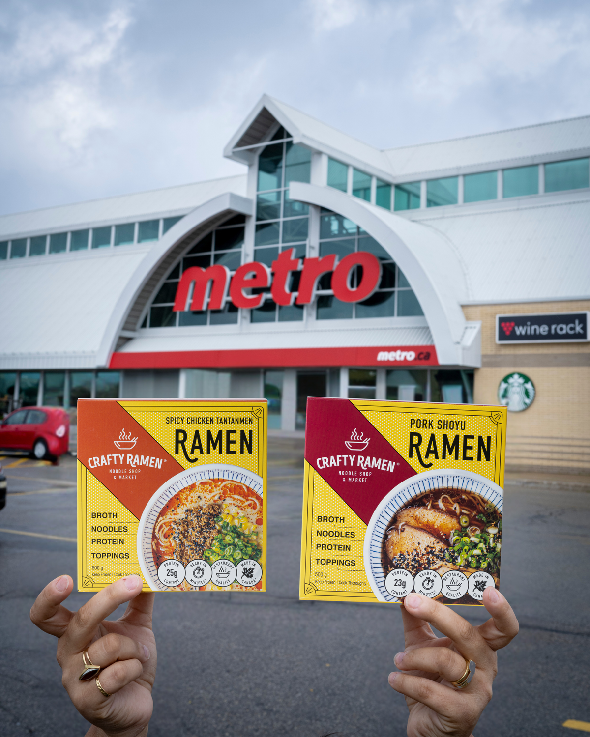 Two hands holding two boxes of Crafty Ramen frozen ramen in front of a Metro store in an outdoor parking lot. The left box is Spicy Chicken Tantanmen Frozen Ramen and the right box is Pork Shoyu Frozen Ramen.