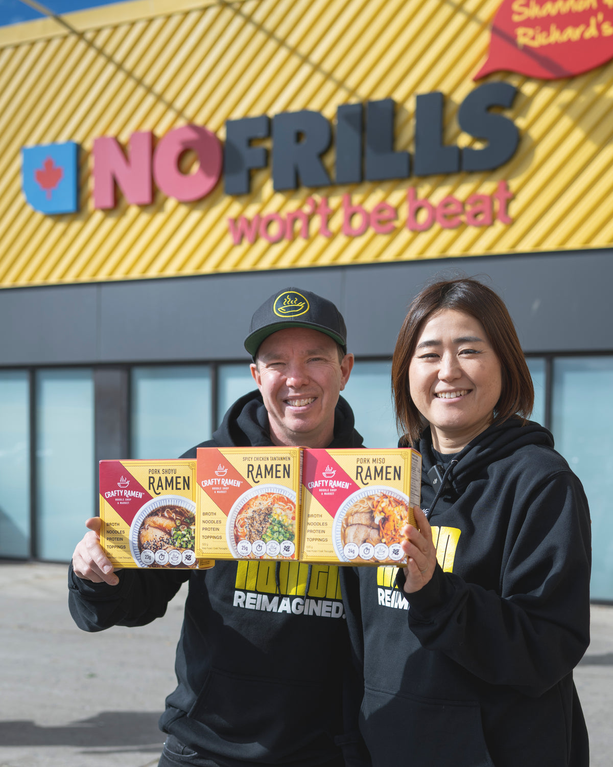 Jared and Miki standing outside in front of a large yellow No Frills sign, holding Crafty Ramen boxes including Spicy Chicken Tantanmen Frozen Ramen, Pork Shoyu Frozen Ramen and Pork Miso Frozen Ramen. Jared on the left is wearing a black Crafty Ramen branded cap and hoodie, and Miki on the right is wearing a black Crafty Ramen hoodie.