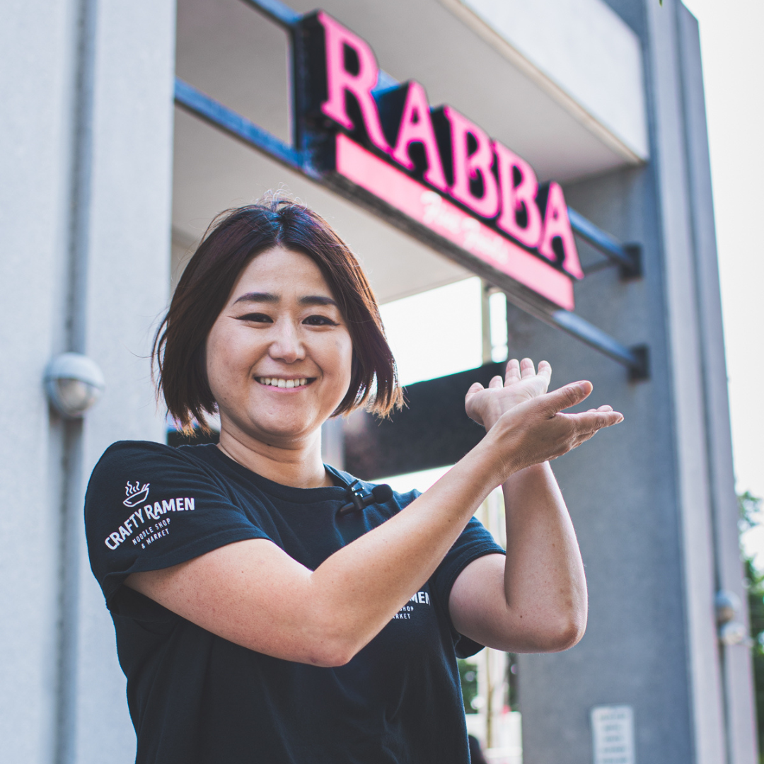 Miki smiling and directing towards a Rabba Fine Foods sign, wearing a black short-sleeve T-shirt with white text that reads 'CRAFT RAMEN' on the sleeve.