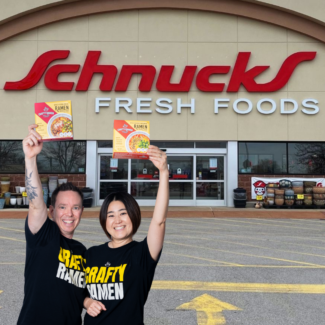 Jared and Miki standing in front of a Schnucks Fresh Foods store, each holding up a box of Crafty Ramen. They are wearing black t-shirts with yellow and white text that reads 'CRAFTY RAMEN'.