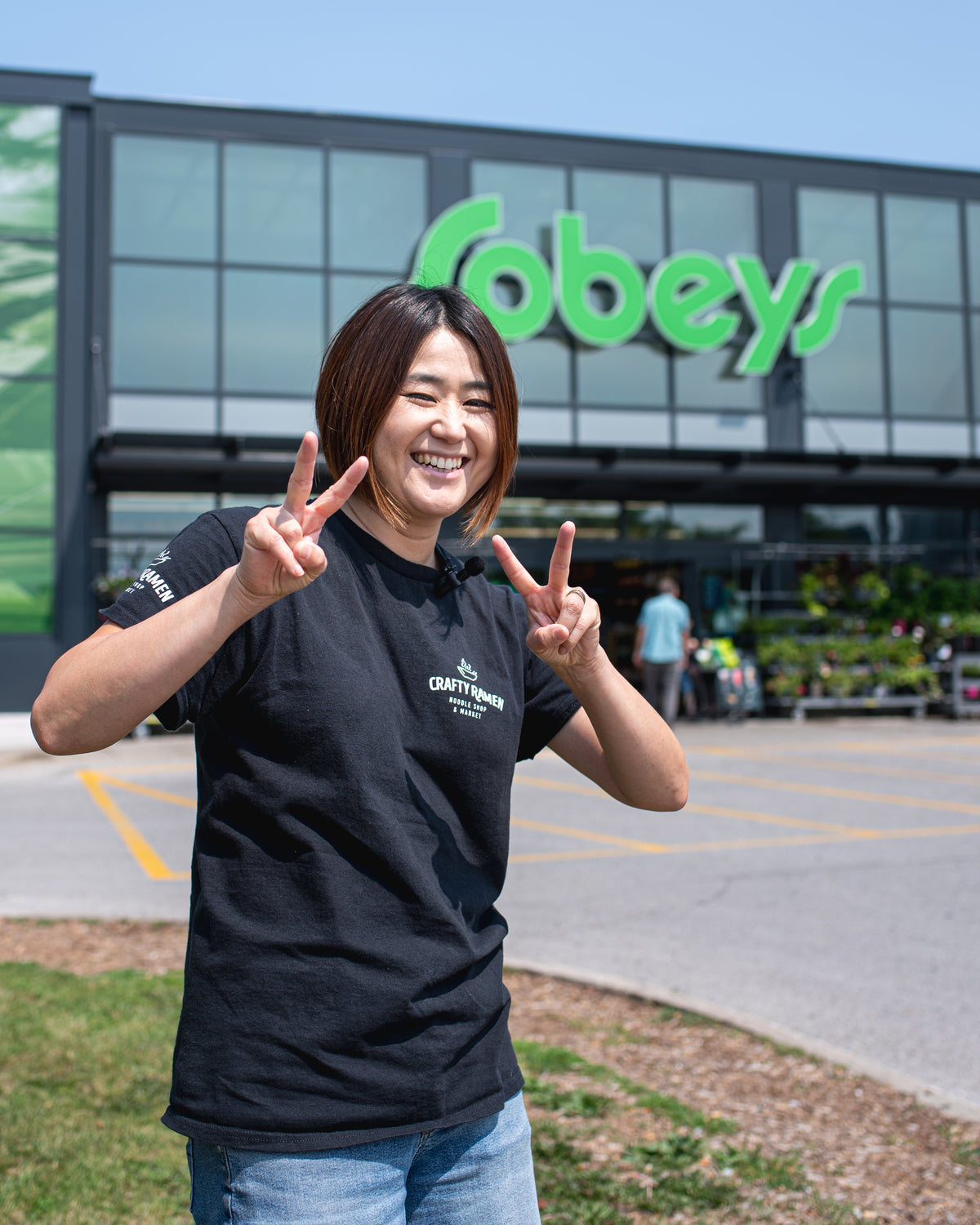 Miki smiling and posing outdoors in front of a Sobeys grocery store, wearing a black crew-neck Crafty Ramen t-shirt with a white printed logo on the chest, and wearing blue jeans.