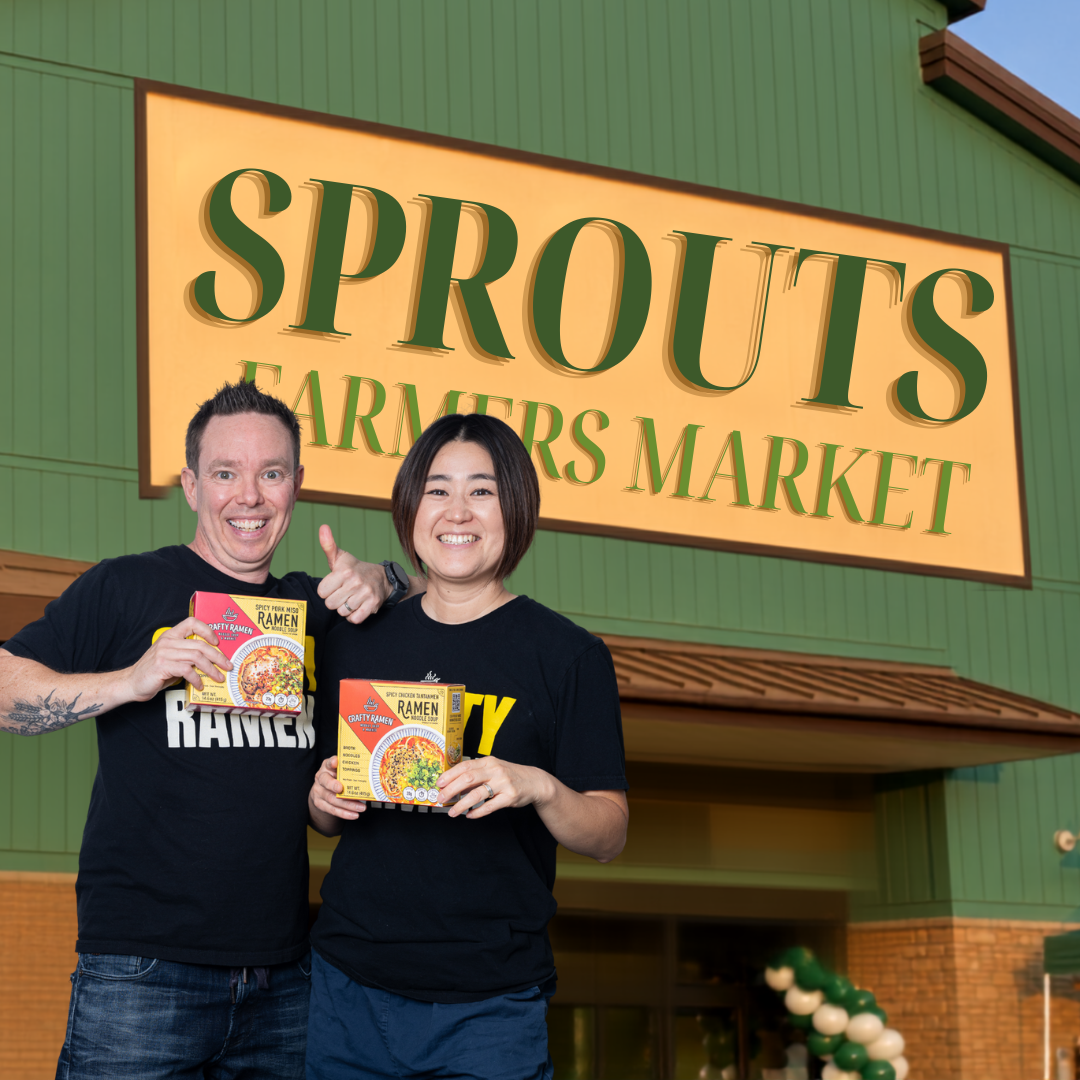 Jared and Miki standing in front of a green building with a large yellow sign reading 'SPROUTS FARMERS MARKET,' each holding a Crafty Ramen box. Both are wearing black T-shirts with white and yellow text that read 'CRAFTY RAMEN'.