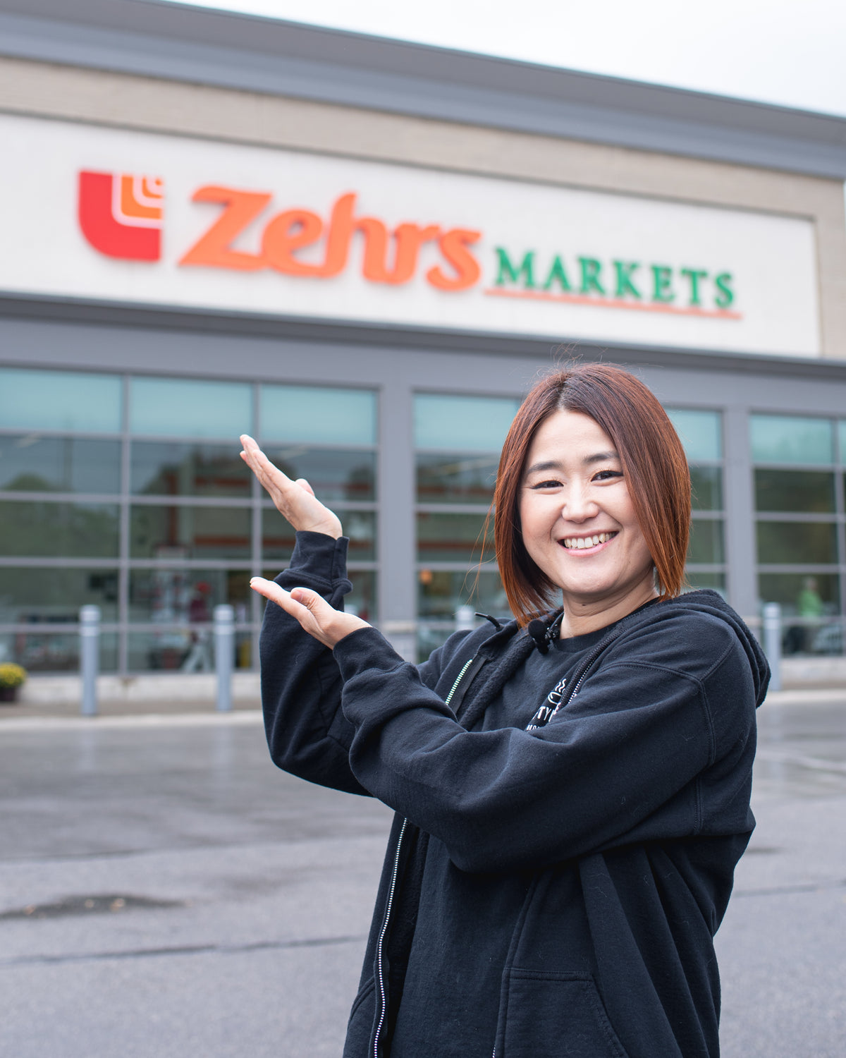 Miki smiling and gesturing with her hands in front of a Zehrs Markets store while wearing a black Crafty Ramen branded t-shirt and hoodie.