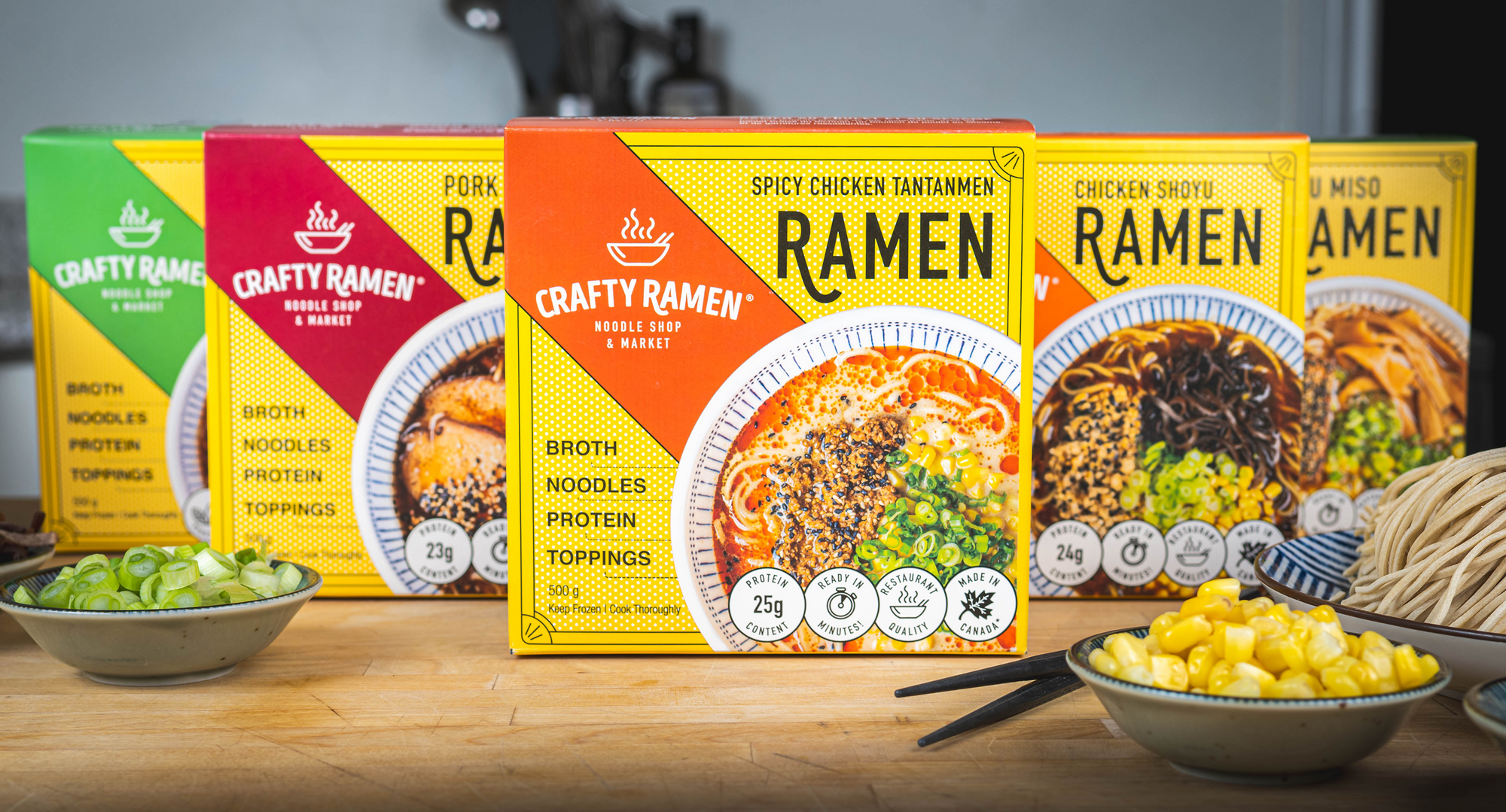 A display of five Crafty Ramen boxes in various flavours (Szechuan Tofu, Pork Shoyu, Spicy Chicken Tantanmen, Chicken Shoyu, and Tofu Miso) with small bowls of corn and green onions in front on a wooden surface.