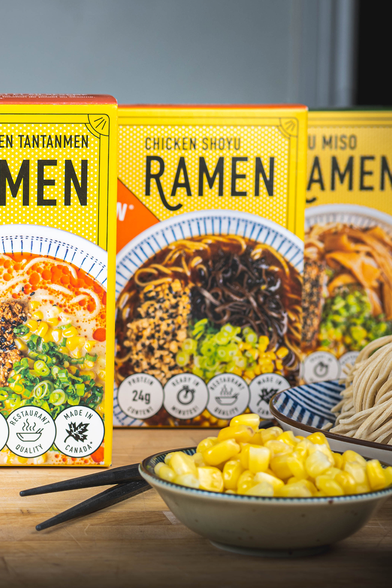 A display of three Crafty Ramen boxes in various flavours (Spicy Chicken Tantanmen, Chicken Shoyu, and Tofu Miso) with a small bowl of corn and chopsticks on a wooden surface.