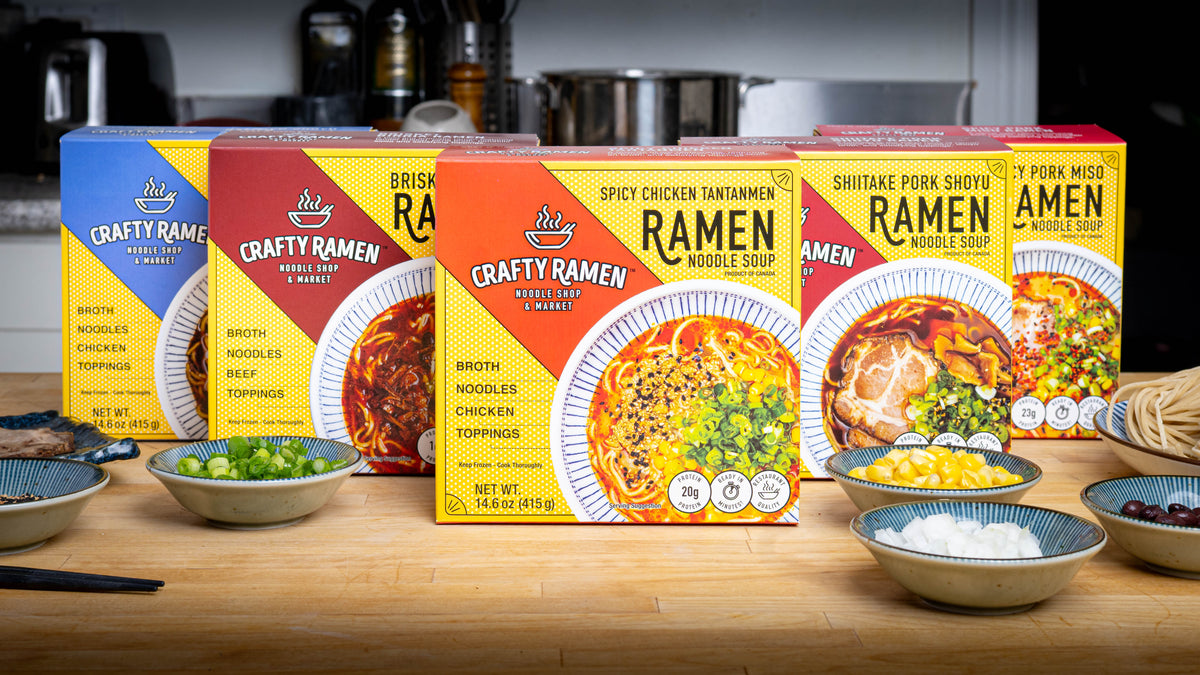 Five boxes of Crafty Ramen Frozen Ramen, including from left to right Chicken Shoyu Frozen Ramen, Brisket Birria Frozen Ramen, Spicy Chicken Tantanmen Frozen Ramen, Shiitake Pork Shoyu Frozen Ramen, and Spicy Pork Miso Frozen Ramen, all lined up on a wooden countertop in a kitchen setting, with small bowls of ingredients such as sweet corn and green onions in front of them.