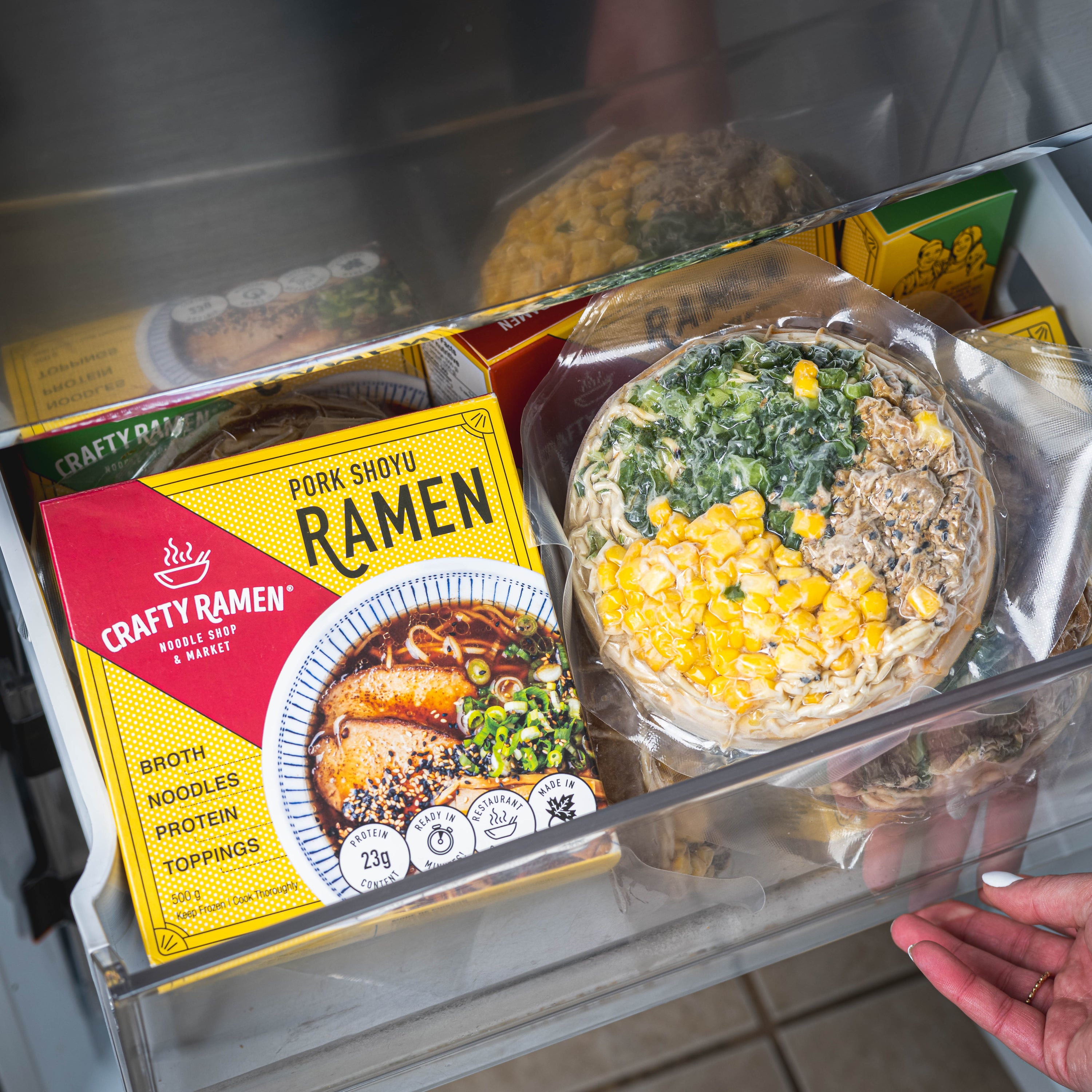 Crafty Ramen Pork Shoyu Frozen Ramen package and Spicy Chicken Tantanmen Frozen Ramen vacuum-sealed puck in a freezer shelf with a hand reaching towards it.