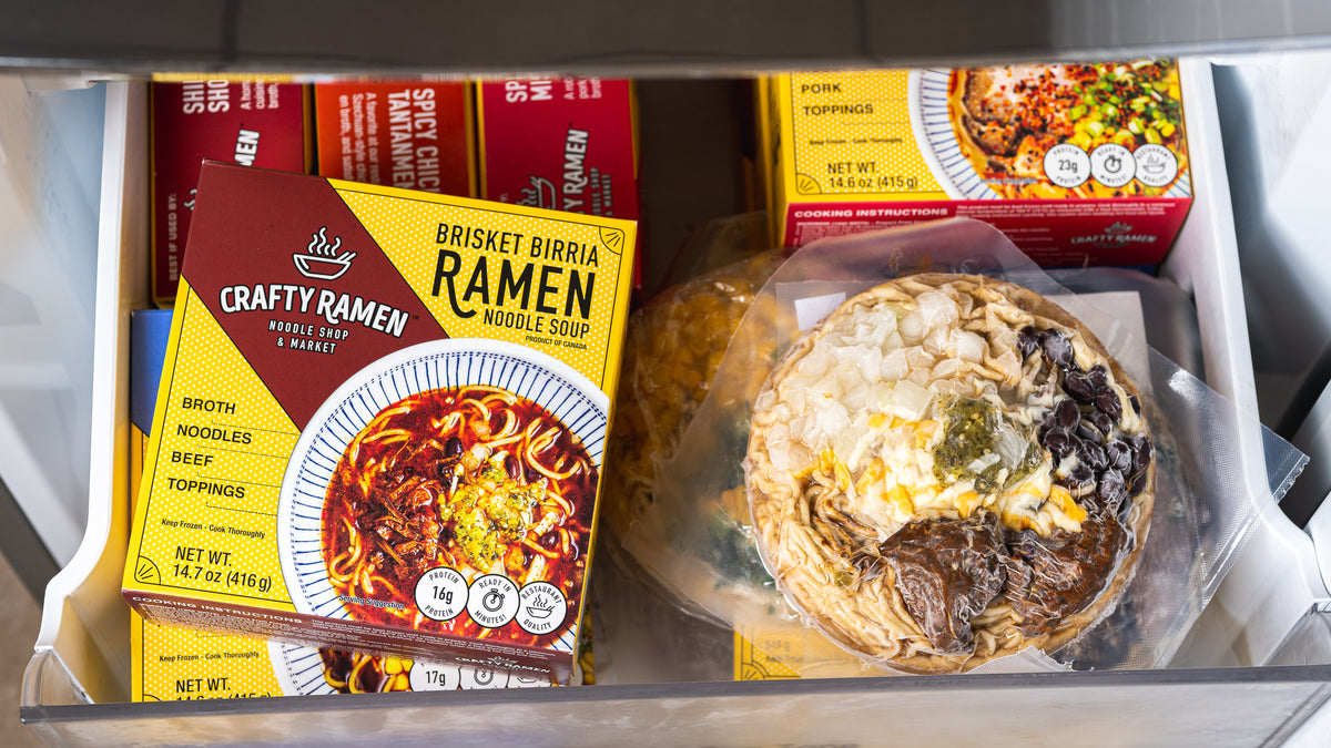 A freezer drawer containing vacuum sealed frozen ramen and Crafty Ramen boxes. The most prominent item is a yellow and red box of Crafty Ramen Brisket Birria Frozen Ramen, with other packaged food items visible behind it.