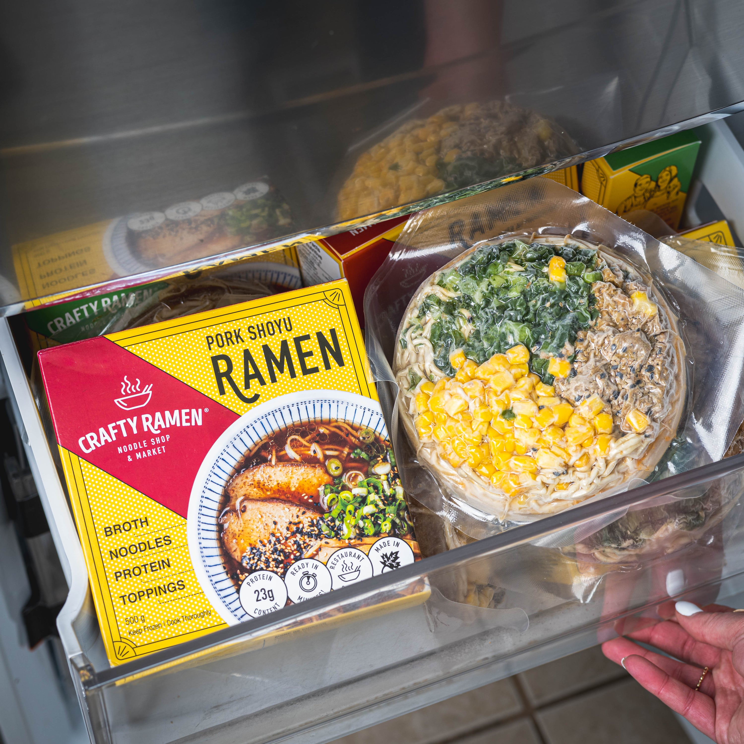 Crafty Ramen Pork Shoyu Frozen Ramen package with a bowl of Spicy Chicken Tantanmen Frozen Ramen in a bottom shelf freezer.