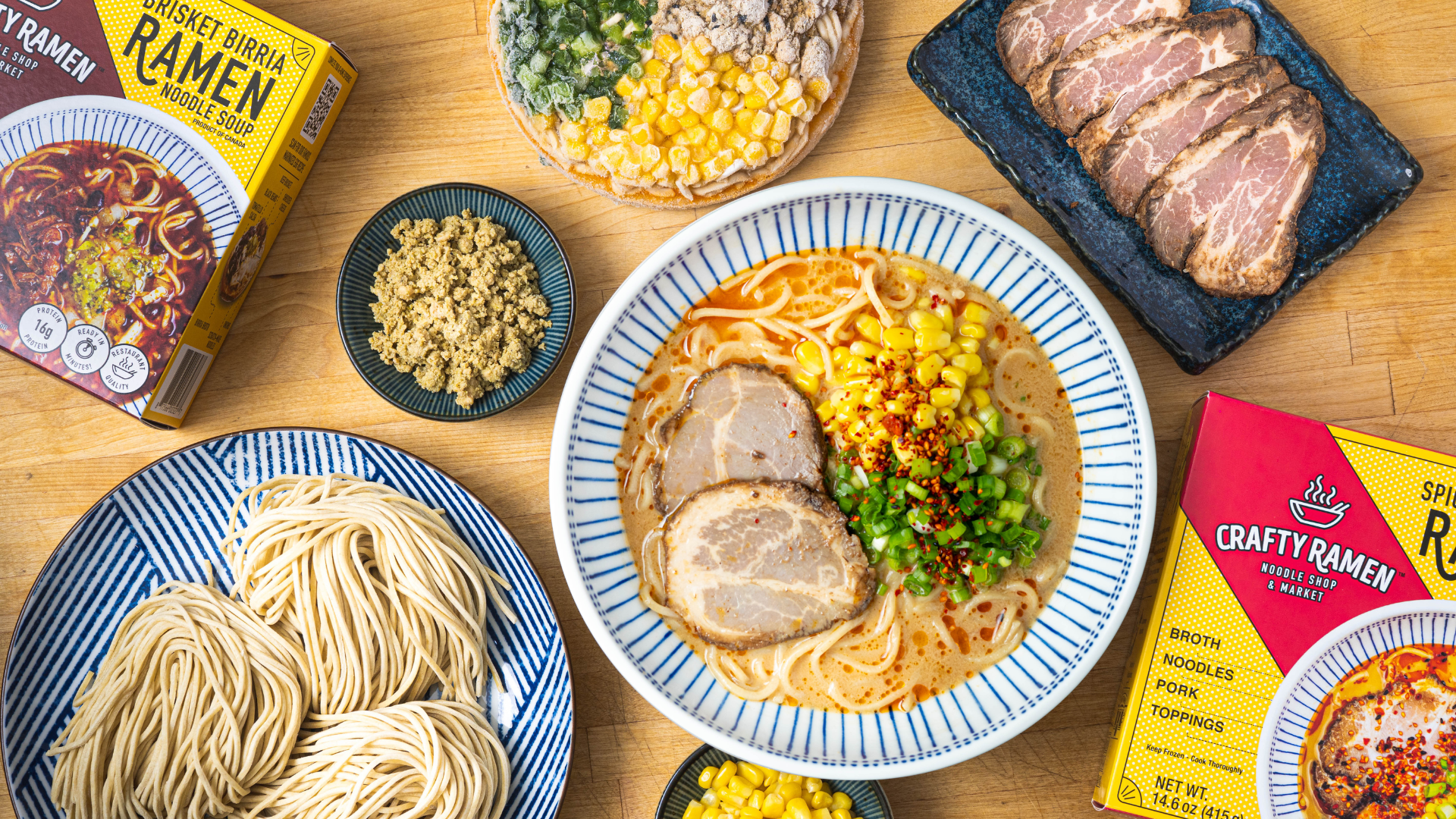 A styled overhead with ingredients and packaged ramen on a wooden table, including a bowl of prepared Spicy Pork Miso Frozen Ramen with sliced pork chashu, scratch-made noodles, sweet corn, green onions, chili crunch, a box of Crafty Ramen Spicy Pork Miso Ramen, and a box of Brisket Birria Frozen Ramen.