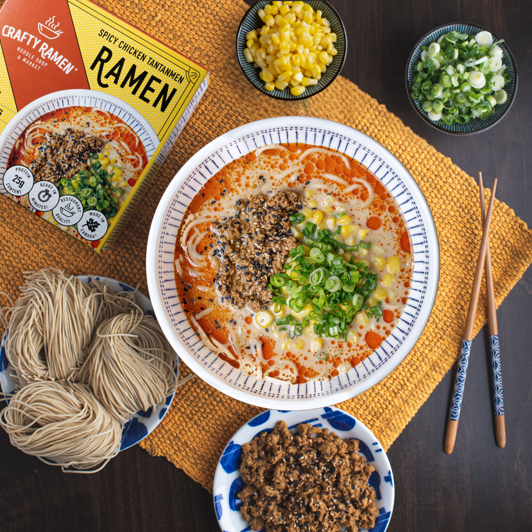 Crafty Ramen Spicy Chicken Tantanmen Frozen Ramen finished bowl with scratch-made noodles, real bone broth, ground sesame chicken, sweet corn, and green onions in a white ramen bowl on a table. Next to it are small bowls of garnishes including ground sesame chicken, sweet corn and green onions, as well as scratch-made noodles on a plate, chopsticks, and a Spicy Chicken Tantanmen Frozen Ramen box.