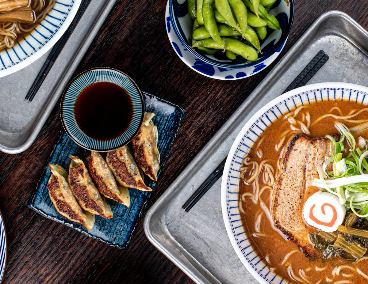 Overhead view of a ramen meal with dumplings, soy dipping sauce, edamame, and a bowl of miso ramen with sliced pork chashu, green onions, and narutomaki on a dark wooden table.