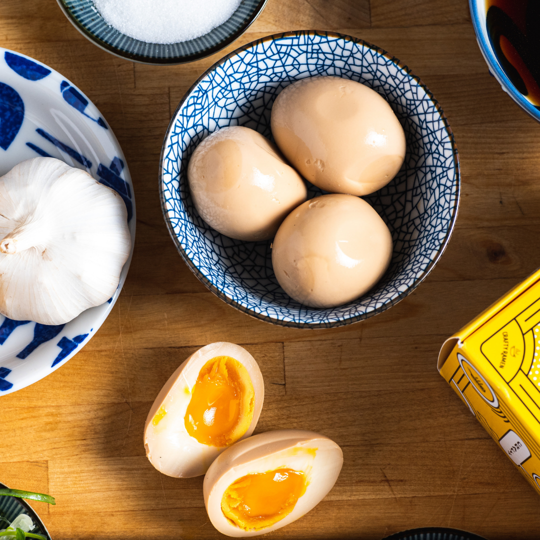 A bowl of soy-marinated eggs next to a yolky soft-boiled egg cut in half on a wooden table.