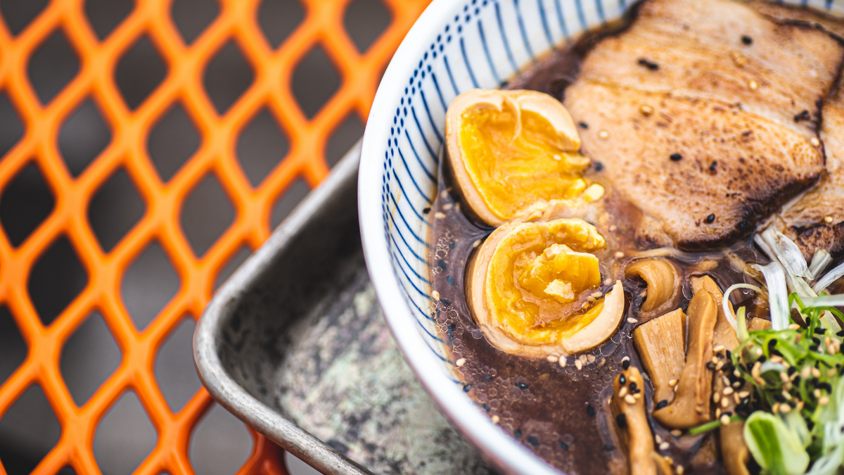 A bowl of ramen with shoyu broth, sliced pork chashu, bamboo shoots, green onions, and two halves of a yolky soft-boiled soy-marined egg on a metal tray.