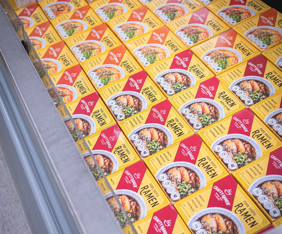 A flat lay in a grocery store displaying multiple yellow boxes of Crafty Ramen products arranged in a grid pattern, each showing a finished bowl of ramen.