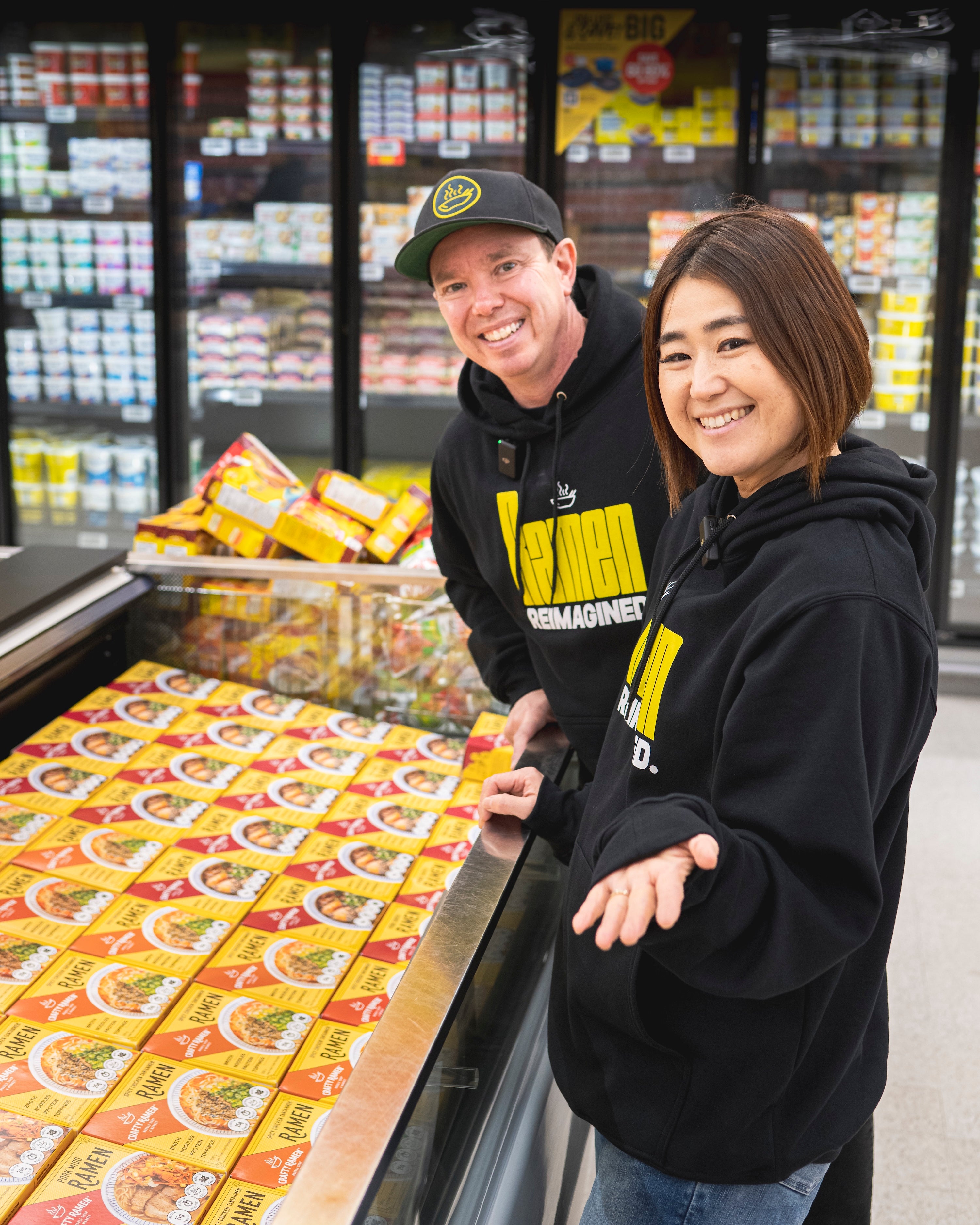 Jared and Miki smiling in front of a bunker full of Crafty Ramen frozen ramen in the frozen aisle of the grocery store.