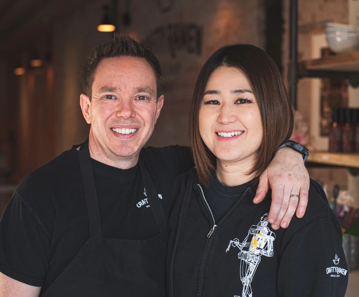 Creators of Crafty Ramen, Jared & Miki, smiling in black Crafty branded clothing standing together indoors in front of a blurred restaurant interior.