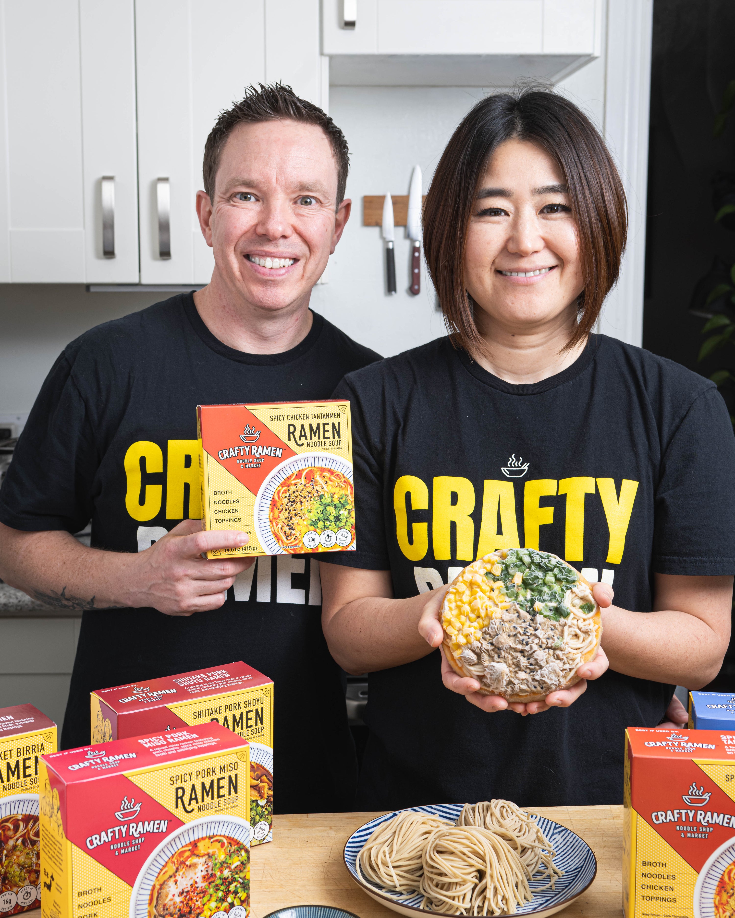 Jared and Miki holding a Spicy Chicken Tantanmen Frozen Ramen box and frozen puck, with the other flavours on a wooden table and scratch-made noodles on a plate.
