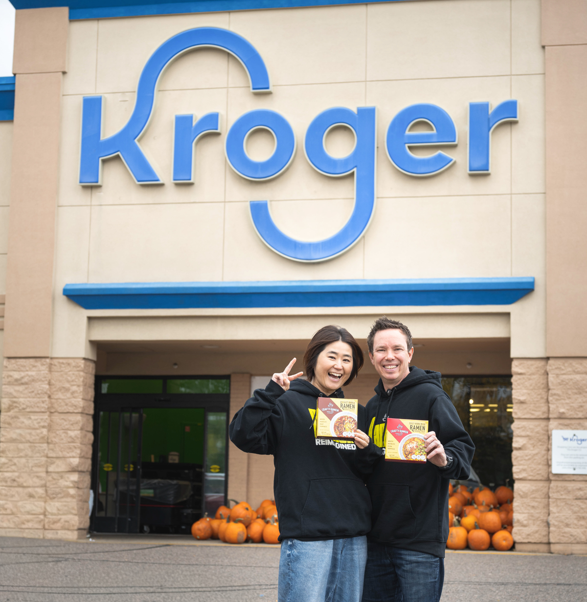 Jared and Miki standing outside of a Kroger holding two boxes of Crafty Ramen.