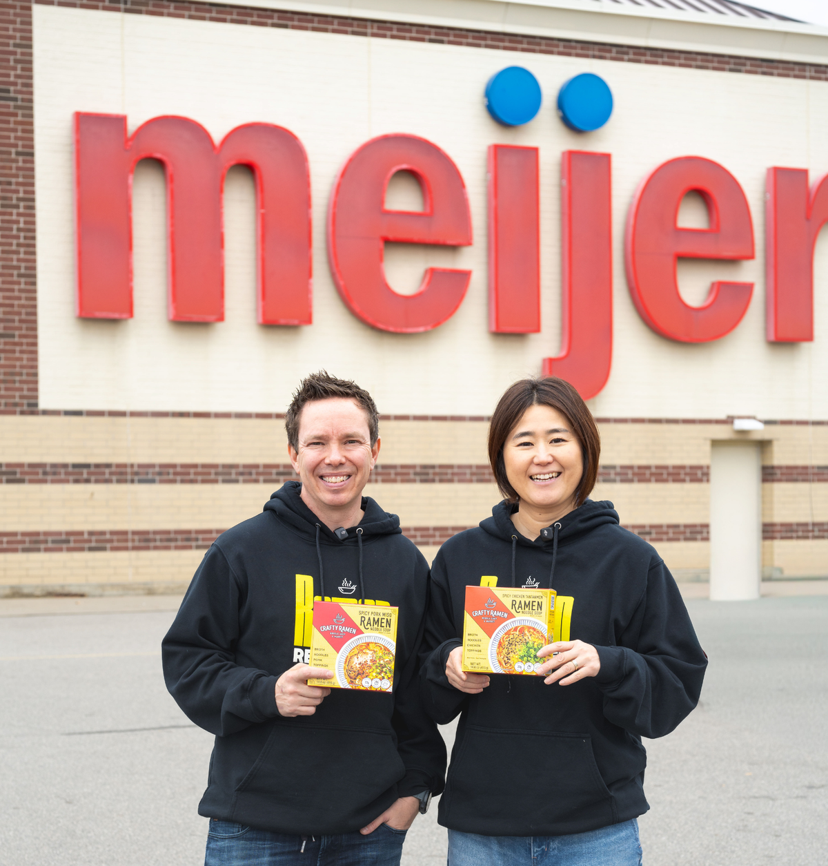 Jared and Miki standing outside of a Meijer holding two boxes of Crafty Ramen.