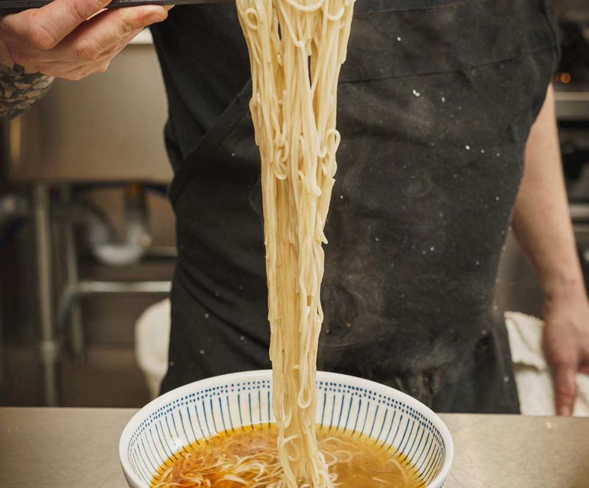 A chef lifting long strands of ramen noodles from a bowl of golden broth using chopsticks, wearing black attire.