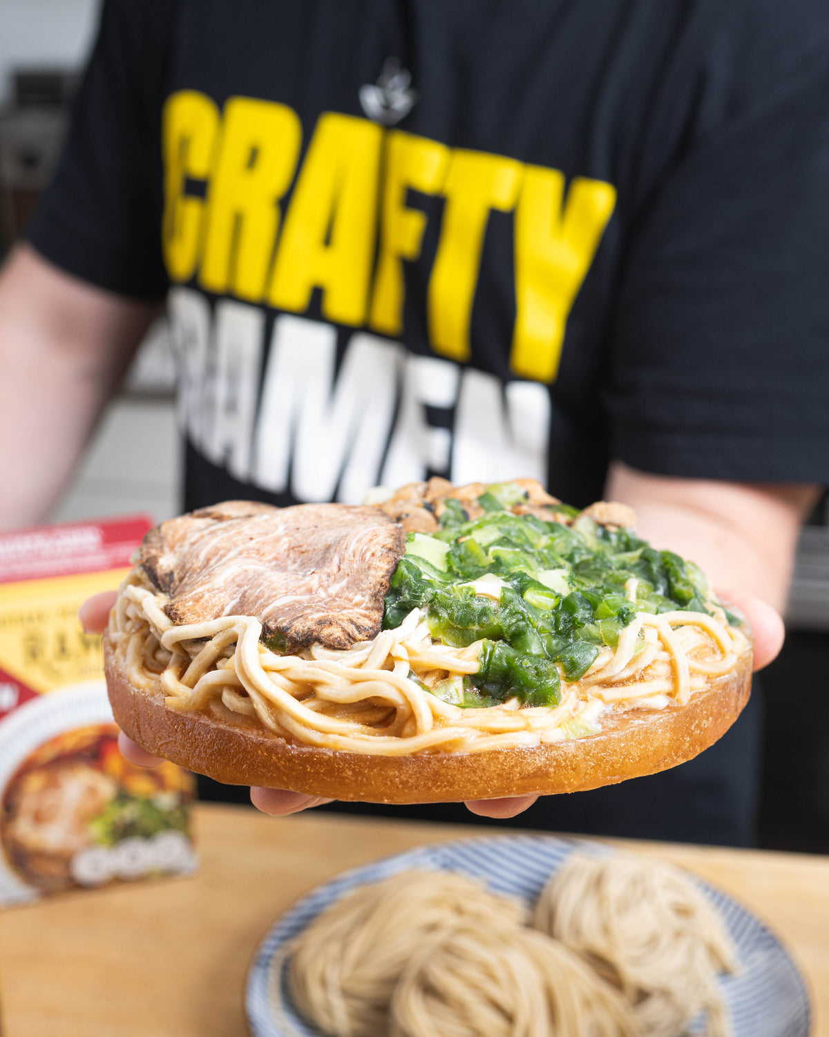 Jared holding a Pork Shoyu Frozen Ramen puck with scratch-made noodles, real bone broth, sliced pork chashu, and green onions, wearing a 'Crafty Ramen' shirt.