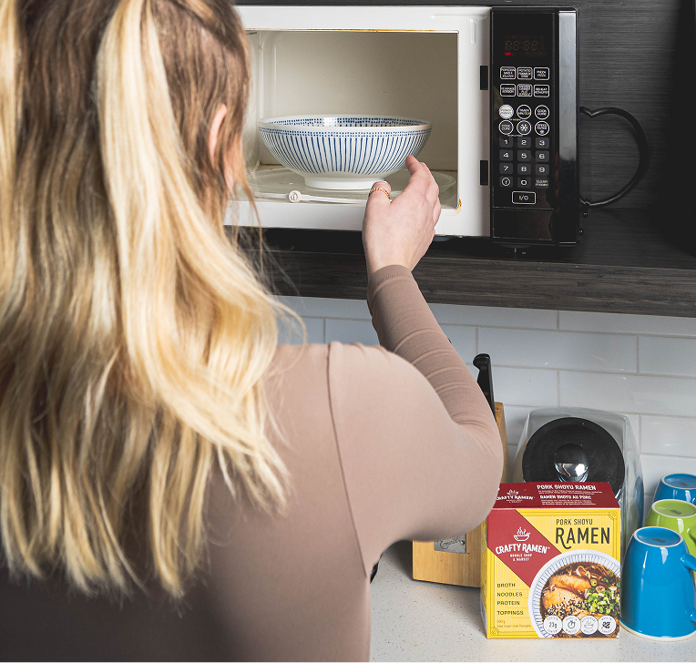 A person placing a blue and white striped ramen bowl into a microwave, with Crafty Ramen packages visible on the shelf below.