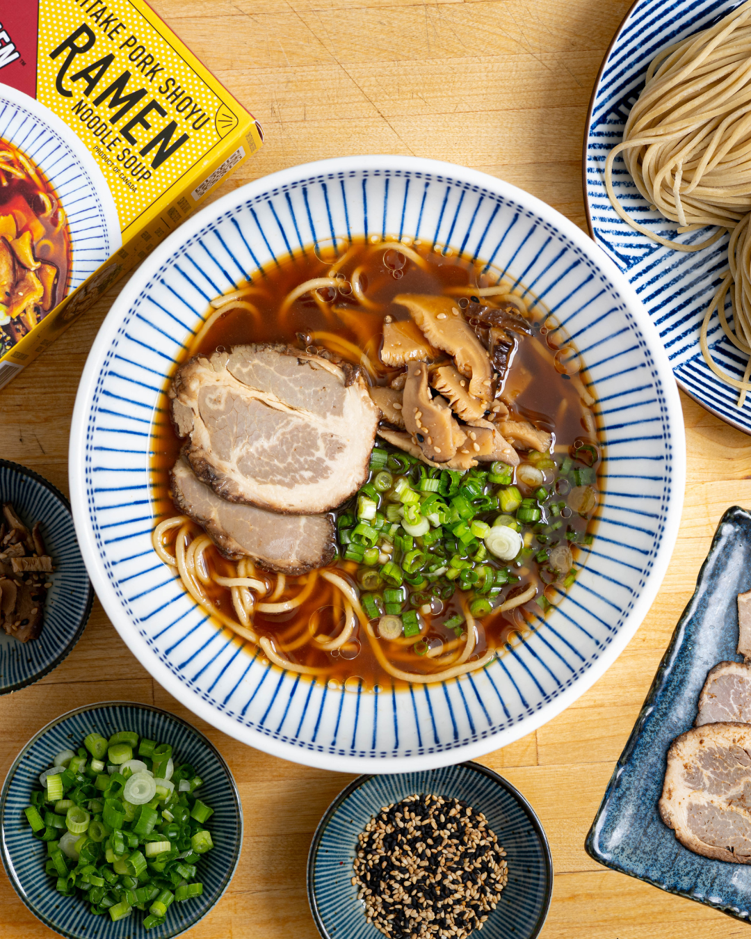 A finished bowl of Shiitake Pork Shoyu Frozen Ramen with sliced pork chashu, green onions, and shiitake mushrooms in a blue-and-white striped bowl, surrounded by small side dishes and a box of Shiitake Pork Shoyu Frozen Ramen on a wooden table.