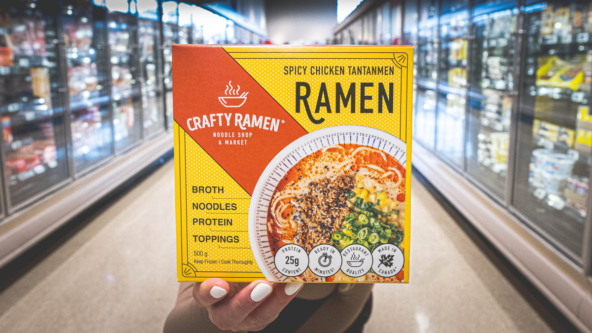 A person holding a box of Crafty Ramen Spicy Chicken Tantanmen Frozen Ramen in a brightly lit grocery aisle with refrigerated shelves in the background.
