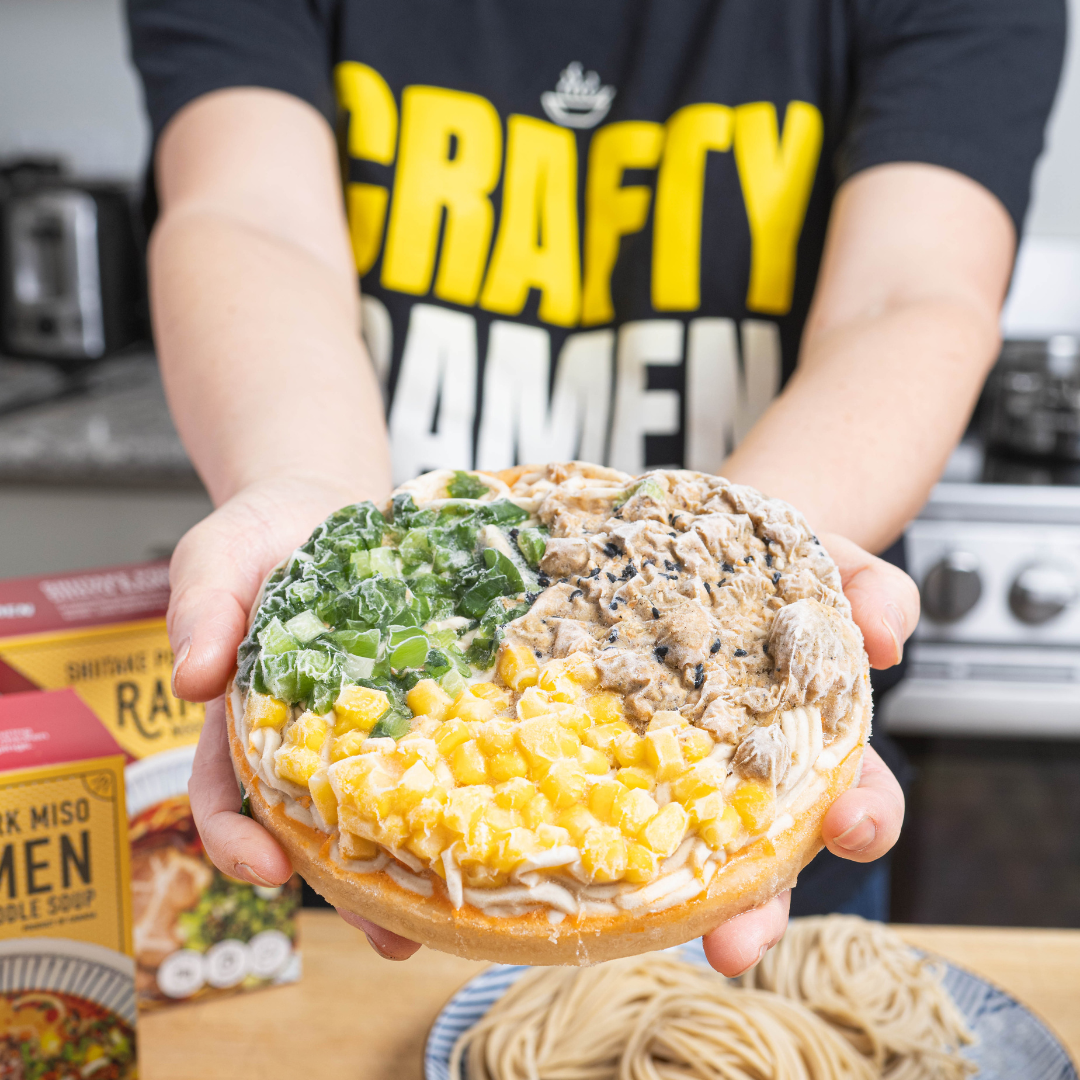Miki in a t-shirt that reads 'Crafty Ramen' printed in yellow holding a Spicy Chicken Tantanmen Frozen Ramen puck with scratch-made noodles, green onions, ground sesame chicken, corn, and real bone broth, with scratch-made noodles in the background.