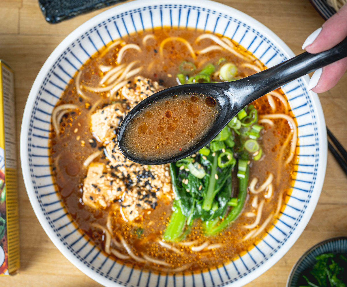 A hand holding a spoon scooping vegan broth from a bowl of Szechuan Tofu Ramen with scratch-made noodles, mapo tofu, sesame seeds, chinese broccoli, and green onions, with a Crafty Ramen box beside it.