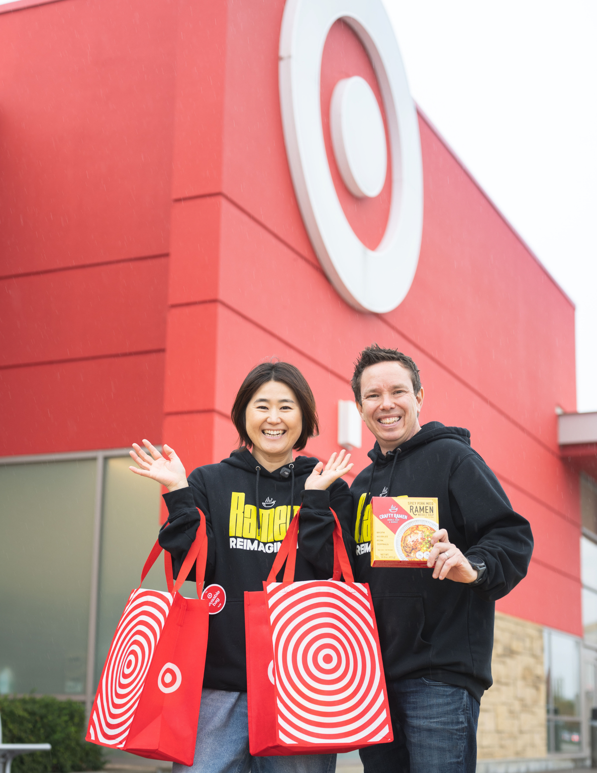 Jared and Miki standing outside of Target holding two Target bags and a Crafty Ramen box wearing Crafty Ramen hoodies with a white bullseye marking the building.