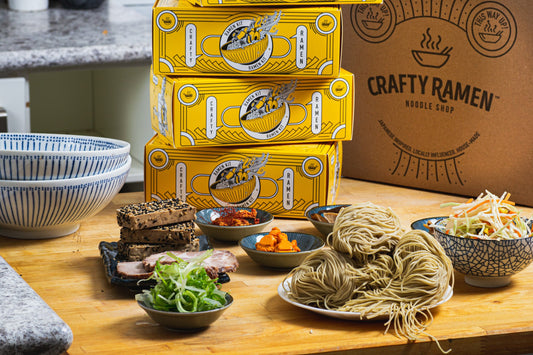 A stack of ramen kits with ingredients mise en place in the foreground.